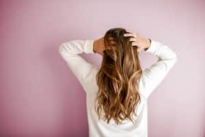 Femme de dos avec les mains sur la tête, cheveux longs, blonds et bouclés, avec fond mur rose