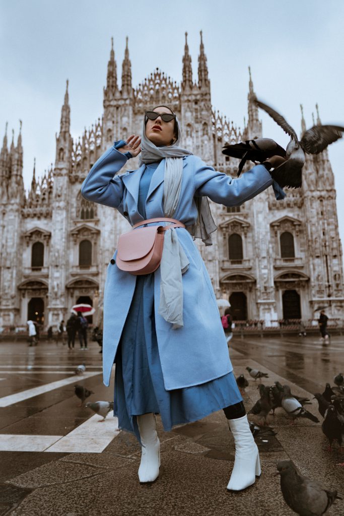 Femme qui porte une robe bleu foncé, un long manteau bleu ciel et un foulard bleu gris, en plus d'un sac banane rose pastel