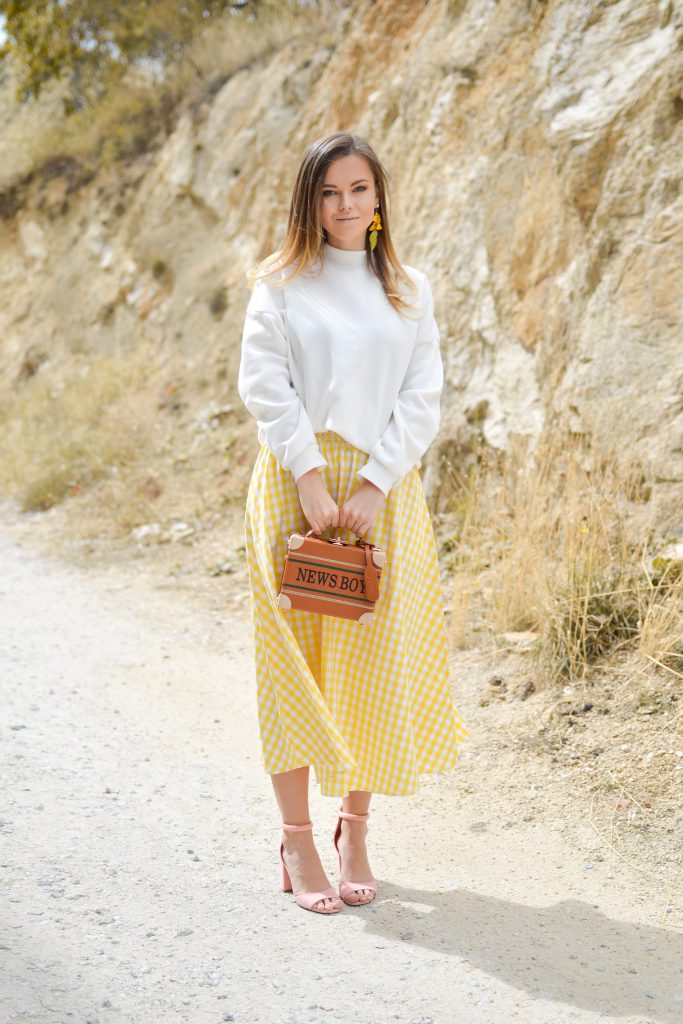 Femme qui porte une jupe jaune clair avec un pull blanc et des sandales roses pâles, ainsi qu'un sac à main en cuir marron