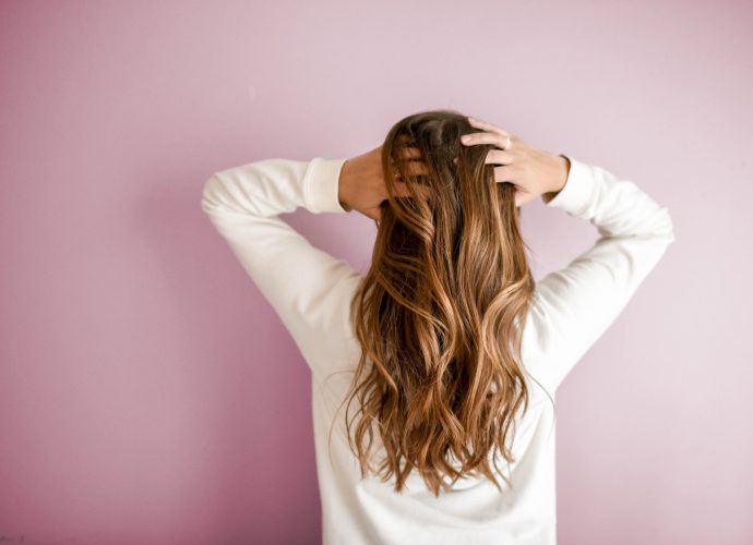 Femme de dos avec les mains sur la tête, cheveux longs, blondset bouclés, avec fond mur rose